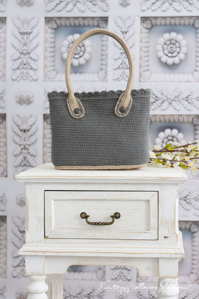 The grey crochet bag sitting on a white end table, with some flowers.