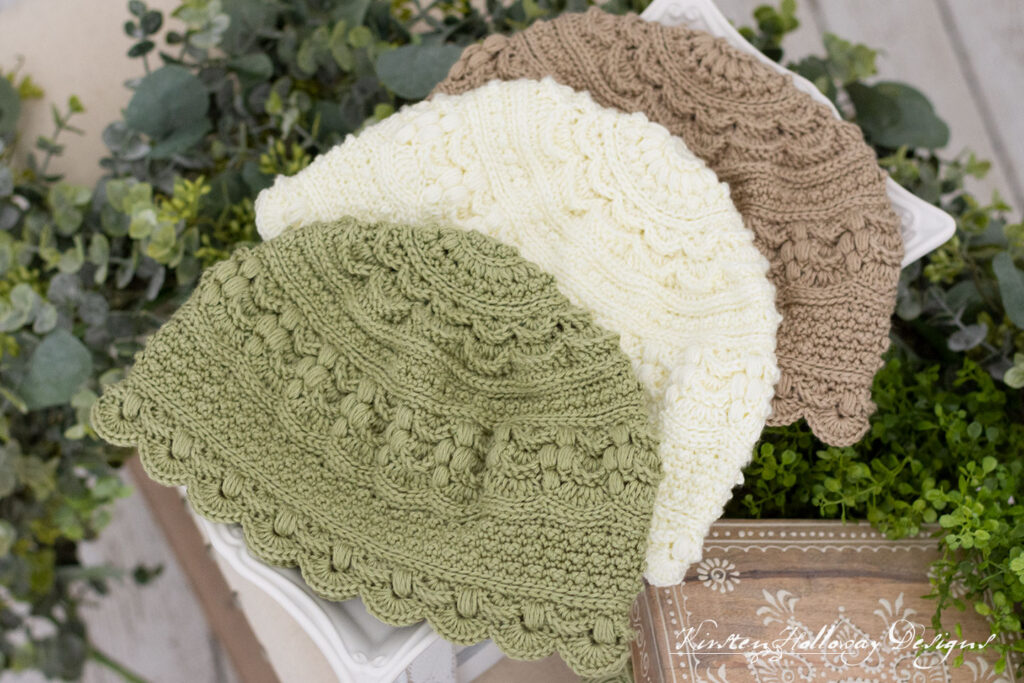 Green, white and light brown crochet hats laying on a serving tray, surrounded by greenery.
