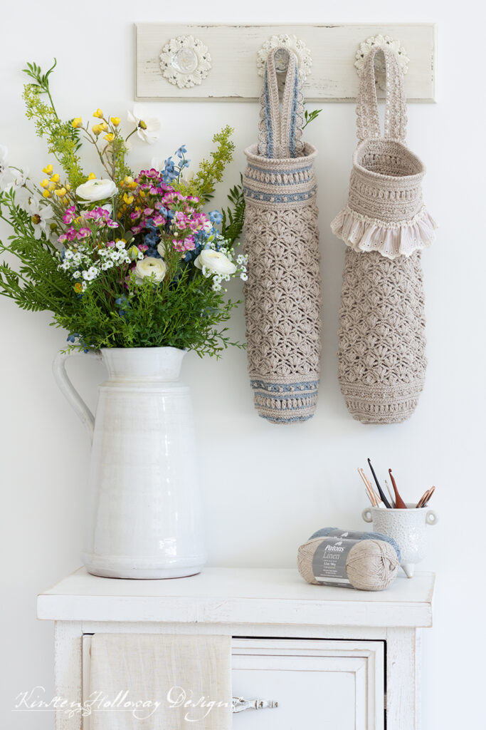 Crochet plastic bag savers in 2 sizes, shown hanging on a white coat rack next to a pot of flowers and above an end table with some skeins of yarn on it.