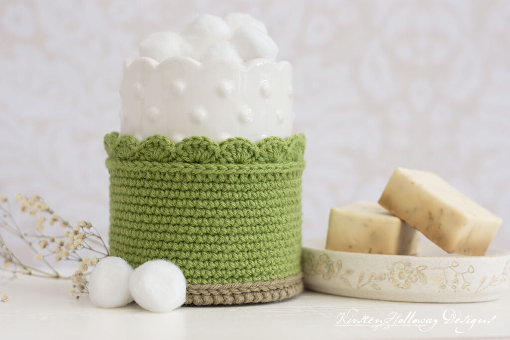 A short, round, fern green crochet basket with some cotton balls in it sitting on a white table. There is a soap dish with 2 small bars of soap next to it, and a couple of cotton balls on the table.